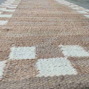 K&ouml;nnte beinhalten: Ein gewebter Teppich mit einem karierten Muster in Beige und Wei&szlig;. Der Teppich ist aus Naturfasern gefertigt und hat einen rustikalen Look.