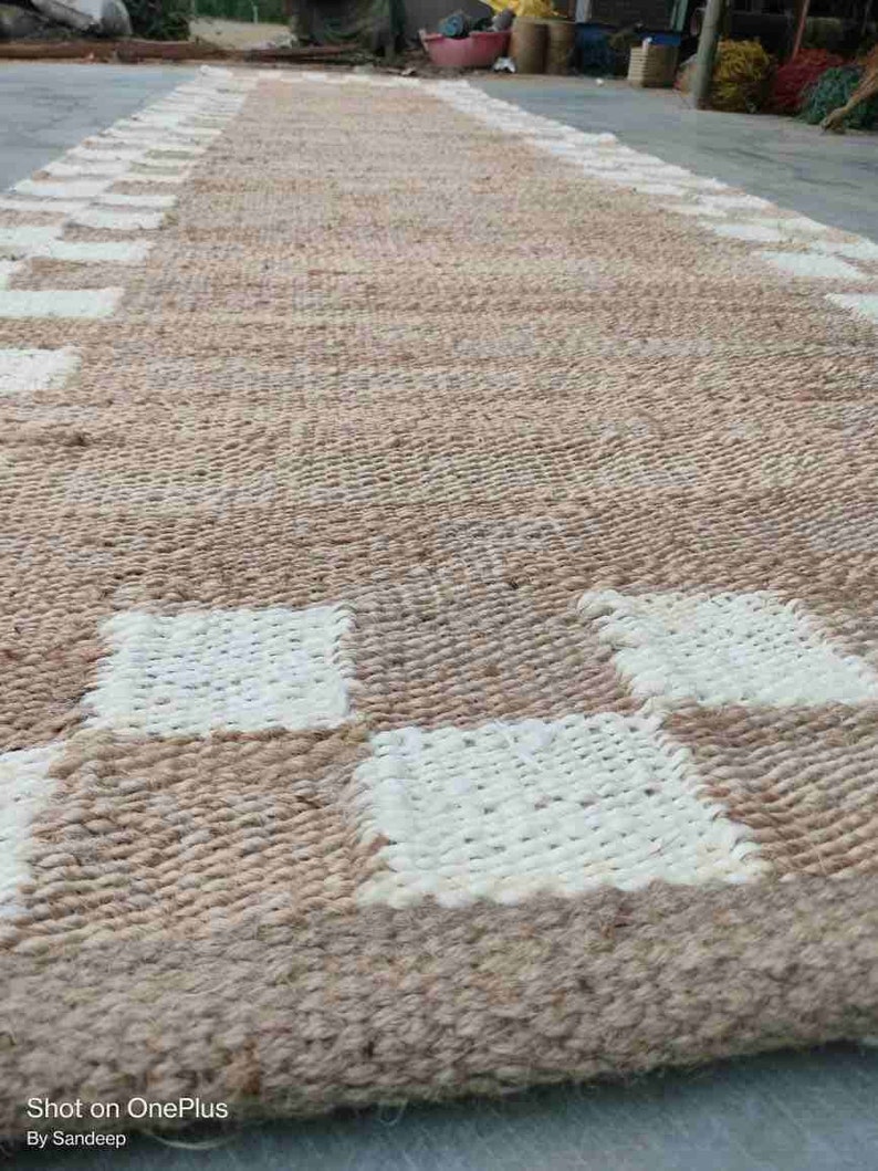 K&ouml;nnte beinhalten: Ein gewebter Teppich mit einem karierten Muster in Beige und Wei&szlig;. Der Teppich ist aus Naturfasern gefertigt und hat einen rustikalen Look.