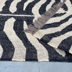 May include: A woven area rug featuring a high-contrast black and off-white zebra stripe pattern. The rug has a folded corner revealing a textured underside and a row of short, dark brown fringe along the edge of the folded section.