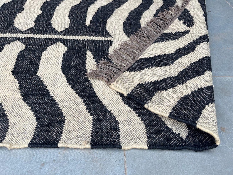 May include: A woven area rug featuring a high-contrast black and off-white zebra stripe pattern. The rug has a folded corner revealing a textured underside and a row of short, dark brown fringe along the edge of the folded section.