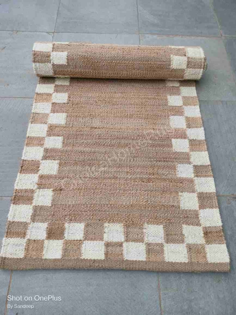 K&ouml;nnte beinhalten: Ein brauner und wei&szlig;er karierter Jute-Teppich mit gewebtem Muster. Der Teppich ist aufgerollt und hat einen Fransenrand.