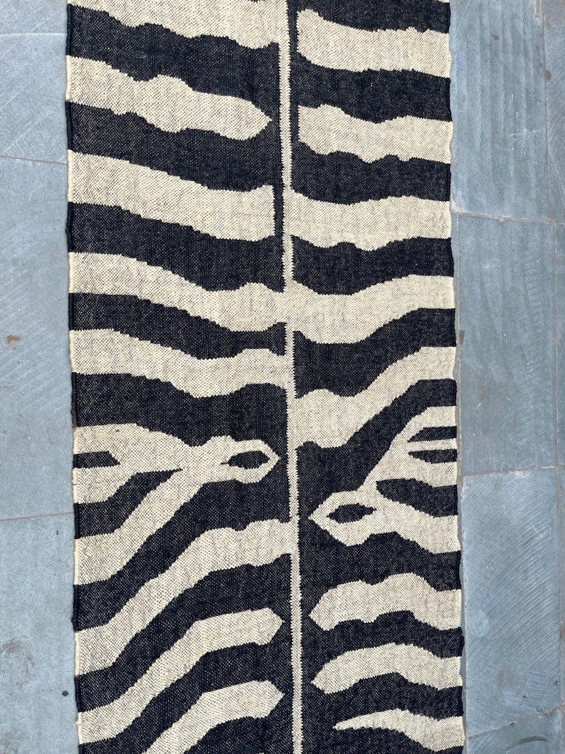 May include: A rectangular woven textile runner featuring a high-contrast black and cream zebra stripe pattern. The design consists of organic, irregular horizontal bands that mirror each other across a central vertical seam, creating a symmetrical look.