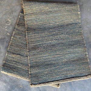 Pode incluir: Um tapete retangular dobrado com uma textura tecida. O tapete apresenta uma mistura de tons azuis e bege, criando um padr&atilde;o listrado. As bordas s&atilde;o finalizadas com uma borda bege, adicionando um contraste sutil ao design geral. Adequado para decora&ccedil;&atilde;o de casa.
