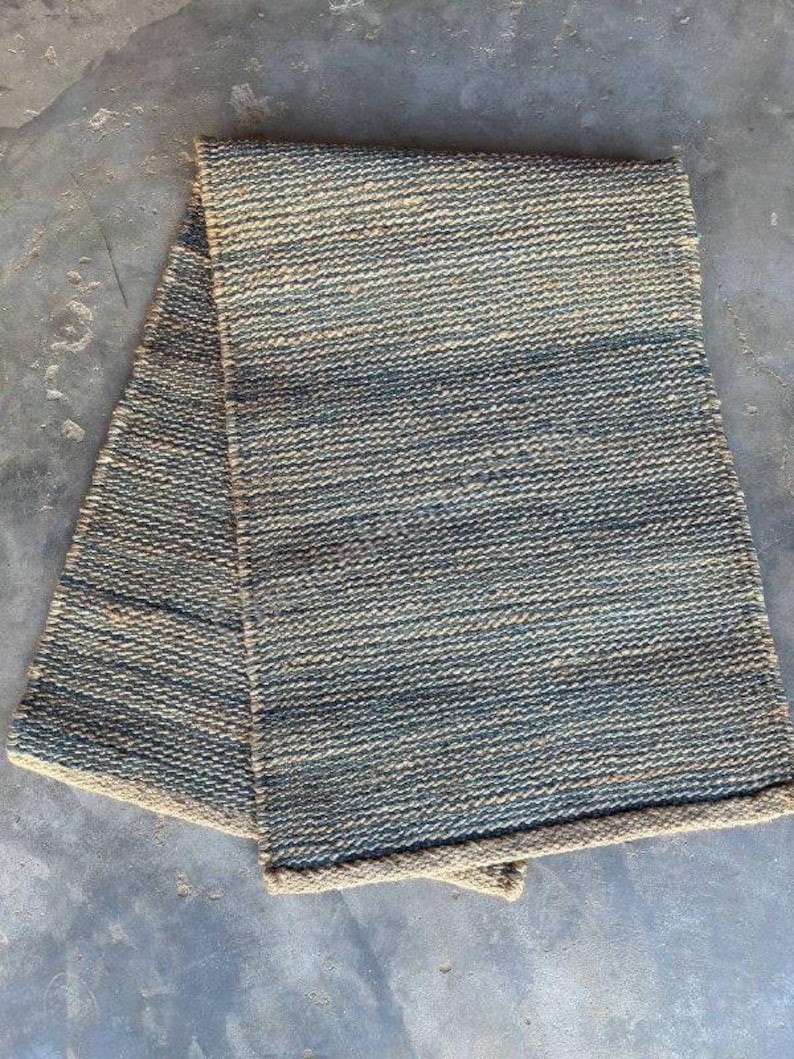 Pode incluir: Um tapete retangular dobrado com uma textura tecida. O tapete apresenta uma mistura de tons azuis e bege, criando um padr&atilde;o listrado. As bordas s&atilde;o finalizadas com uma borda bege, adicionando um contraste sutil ao design geral. Adequado para decora&ccedil;&atilde;o de casa.