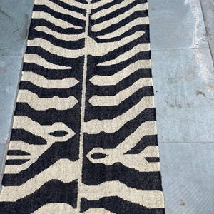 May include: A rectangular woven runner rug featuring a high-contrast black and cream zebra-inspired pattern. The design consists of a central vertical spine with symmetrical, branching horizontal stripes extending outward. The rug has short fringe.