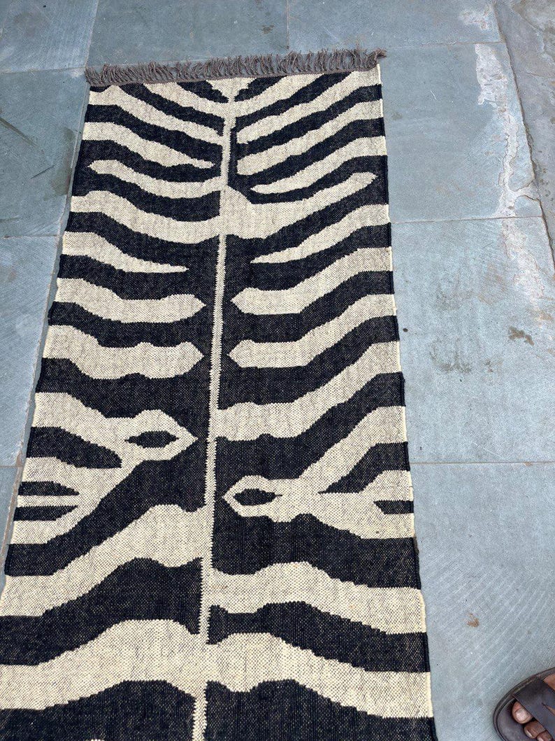 May include: A rectangular woven runner rug featuring a high-contrast black and cream zebra-inspired pattern. The design consists of a central vertical spine with symmetrical, branching horizontal stripes extending outward. The rug has short fringe.