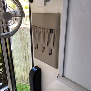 May include: A beige fabric wall organiser with three hooks and a pocket. The organiser is mounted on a white wall.