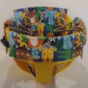 Könnte beinhalten: Ein gelber Babysitz mit einem farbenfrohen Stofffutter mit Zeichentieren wie Löwen, Zebras und Giraffen. Der Sitz hat eine schwarze Schnalle und Metallstützstangen. Der Stoff hat einen blauen Hintergrund mit orangefarbenen, grünen und gelben Akzenten.