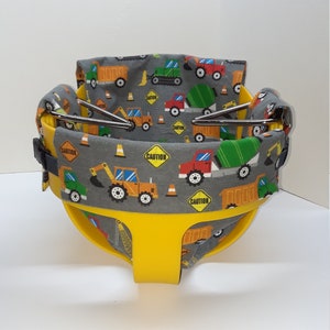 May include: Yellow baby swing seat with a grey fabric cover featuring a construction vehicle print. The fabric has the words "Caution" printed on it.