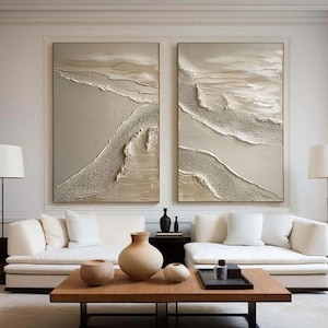 May include: Two abstract paintings in beige and white tones with textured brushstrokes, hanging on a white wall above a white couch and a wooden coffee table.