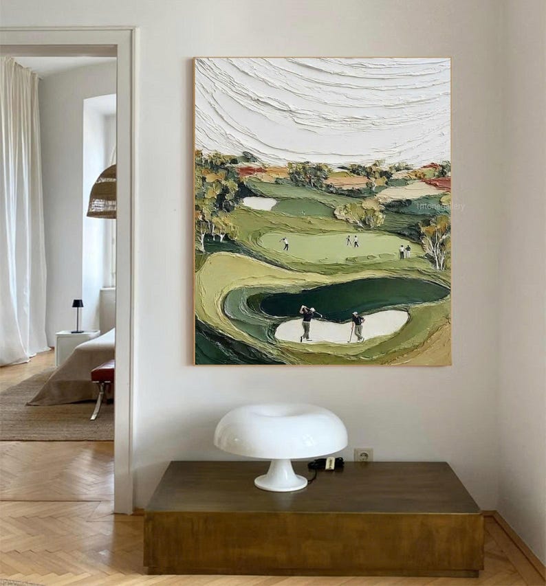 May include: Textured oil painting of a golf course with players. The artwork features a green and tan landscape with white sand traps and a textured white sky. The painting is framed and hangs on a white wall.