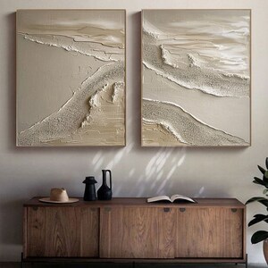 May include: Two abstract paintings with a beige and white color scheme. The paintings feature a textured, layered design with raised lines and swirls. The paintings are framed in a thin, light wood frame.
