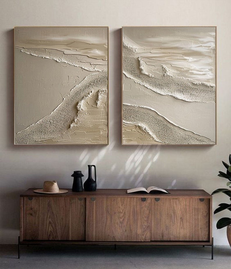 May include: Two abstract paintings with a beige and white color scheme. The paintings feature a textured, layered design with raised lines and swirls. The paintings are framed in a thin, light wood frame.