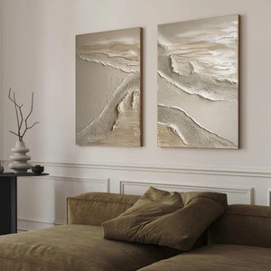 May include: Two abstract paintings with a textured, layered look. The paintings are in shades of beige, brown, and white, and they feature a flowing, organic design. The paintings are hung on a white wall above a brown couch.