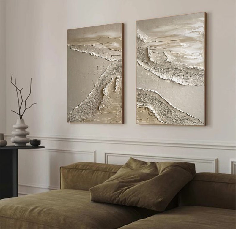May include: Two abstract paintings with a textured, layered look. The paintings are in shades of beige, brown, and white, and they feature a flowing, organic design. The paintings are hung on a white wall above a brown couch.