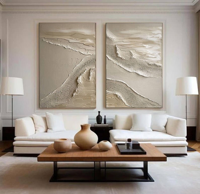May include: Two abstract paintings in beige and white tones with textured brushstrokes, hanging on a white wall above a white couch and a wooden coffee table.