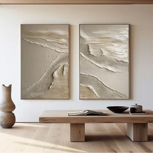 May include: Two abstract paintings with a beige background and white and gold textured lines. The paintings are framed and hanging on a white wall.