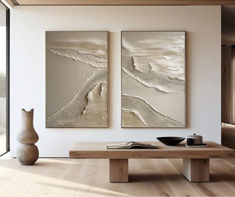 May include: Two abstract paintings with a beige background and white and gold textured lines. The paintings are framed and hanging on a white wall.