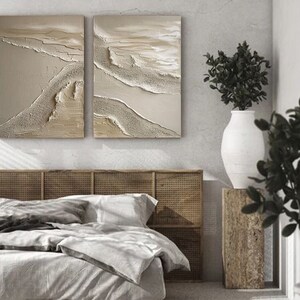 May include: Two abstract paintings with a beige and white color scheme. The paintings are hung on a wall above a bed with a woven headboard.