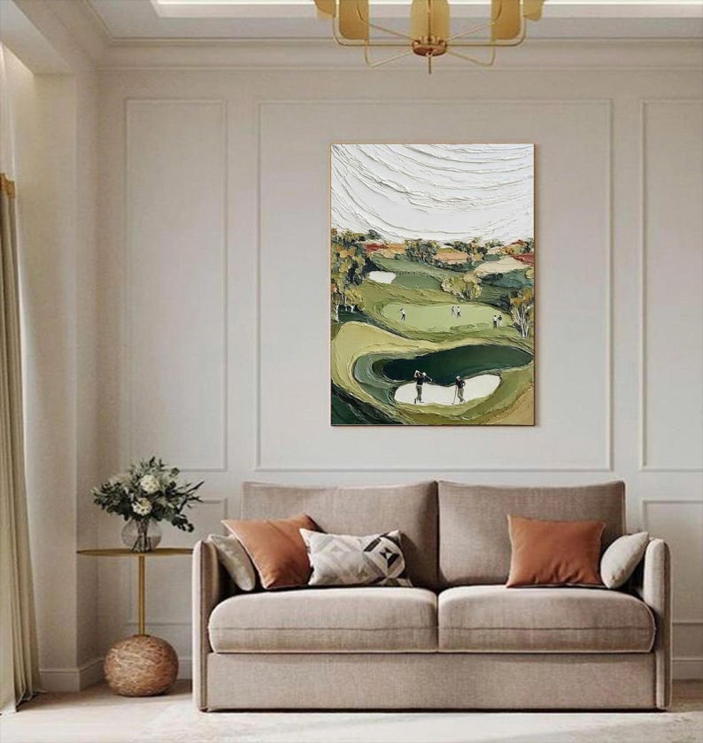 May include: An abstract golf course painting with a textured, layered effect. The artwork features a green golf course with players, sand traps, and trees. The sky is white with textured brushstrokes. The painting is displayed above a beige sofa with orange and patterned pillows.