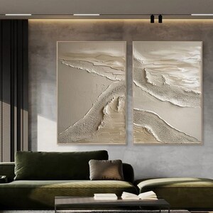 May include: Two abstract paintings with a textured, layered look. The paintings are in shades of beige and white, with a subtle shimmer. They are hung on a gray wall above a green sofa.