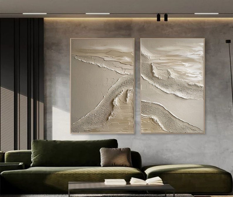 May include: Two abstract paintings with a textured, layered look. The paintings are in shades of beige and white, with a subtle shimmer. They are hung on a gray wall above a green sofa.