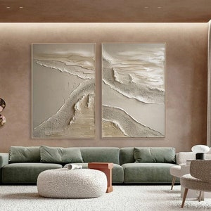 May include: Two abstract paintings in a modern living room. The paintings are both beige and white with a textured, layered look. The paintings are hung on a beige wall above a green sofa.