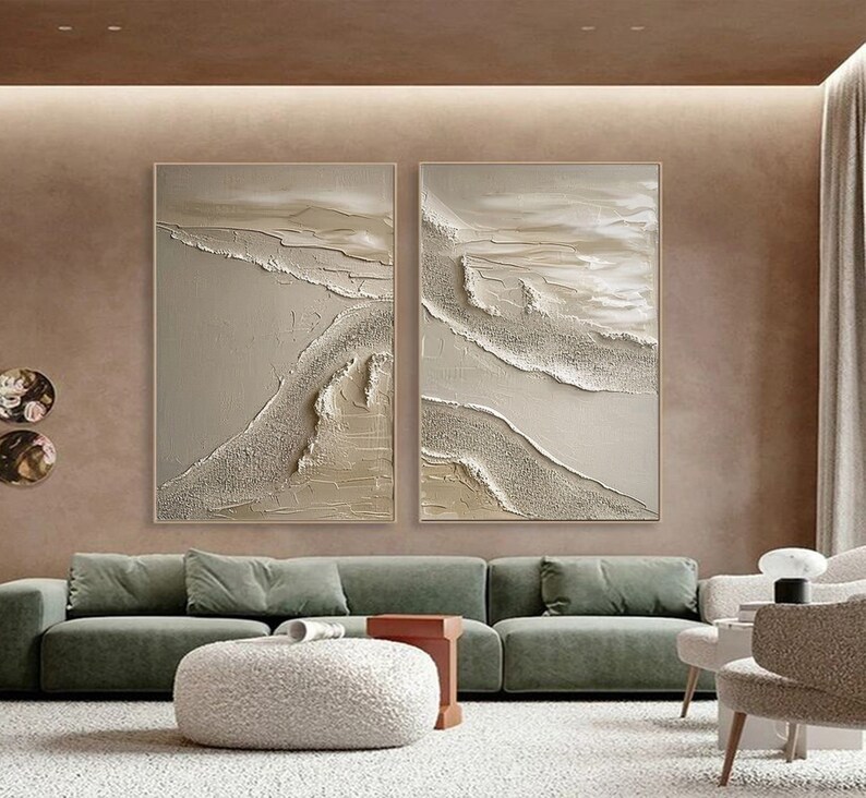 May include: Two abstract paintings in a modern living room. The paintings are both beige and white with a textured, layered look. The paintings are hung on a beige wall above a green sofa.