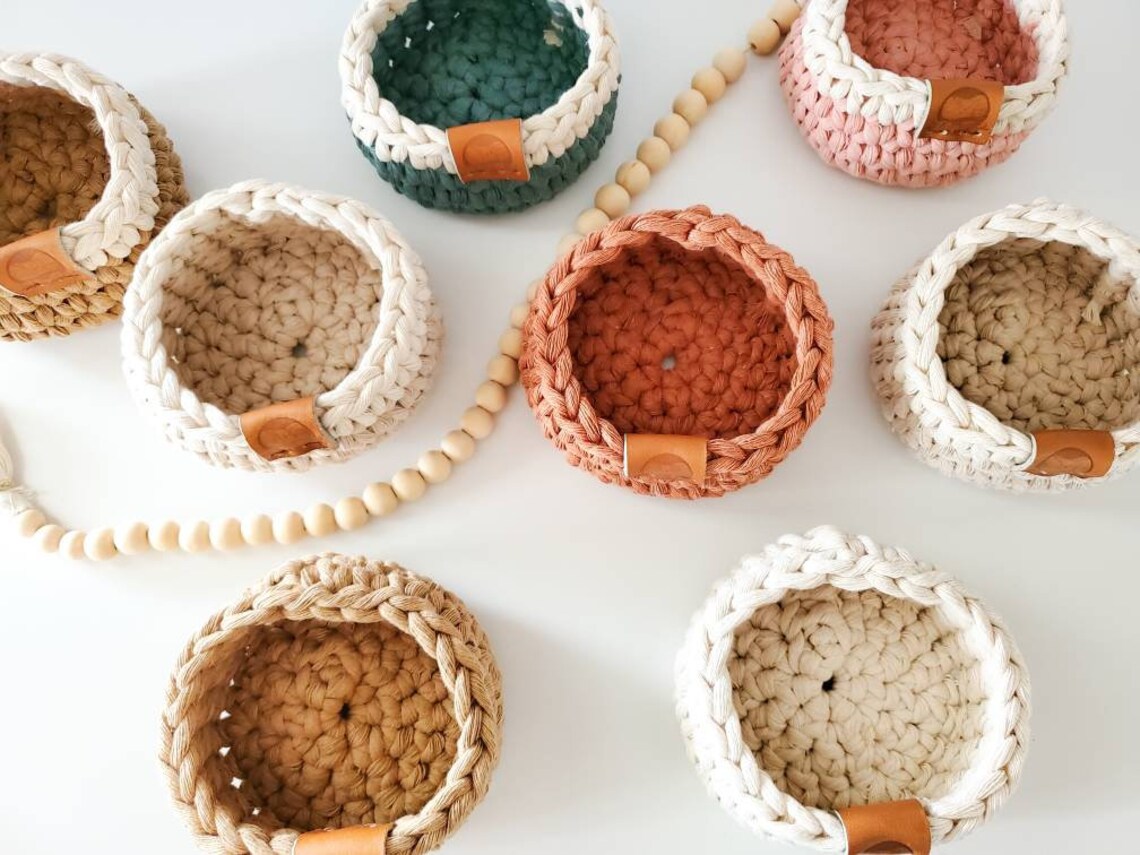 May include: Seven crocheted baskets in various colors including beige, brown, green, and pink. The baskets are all round and have a leather handle. The baskets are arranged on a white surface with a string of wooden beads.