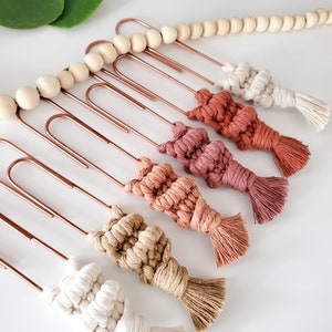 May include: A set of six macrame tassel bookmarks in various shades of brown, beige, and white, hanging from copper-colored paper clips. The tassels are attached to wooden beads.