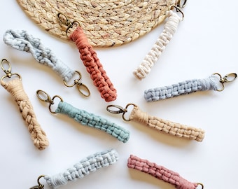 May include: A collection of nine colorful macrame keychains with different colored cords and metal key rings. The keychains are arranged on a white surface with a woven straw mat in the background.