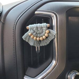 May include: A light blue macrame car air freshener with wooden beads hanging from the bottom. The air freshener is attached to a car's air vent.