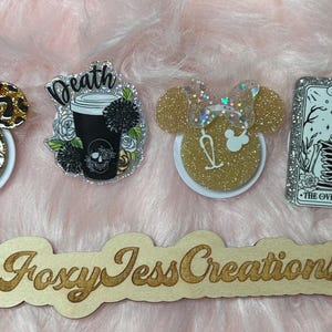May include: Four phone grips with different designs. One is a gold glitter Minnie Mouse head, one is a leopard print Minnie Mouse head with a red bow, one is a black and white coffee cup with the word "Death" above it, and one is a tarot card with a skeleton and the words "The Overthinker" on it.
