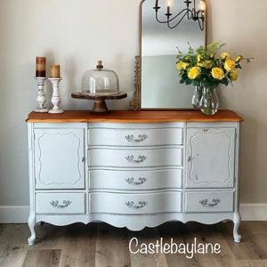 May include: A light gray painted antique dresser with a wood top and ornate silver hardware. The dresser has six drawers and two doors. The dresser is sitting on a wood floor. The dresser is decorated with a glass cloche and candles.