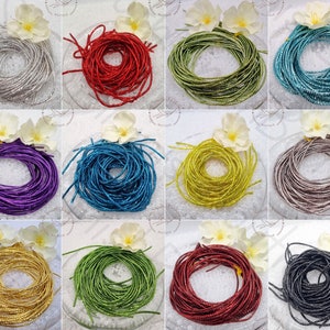 Op de afbeelding: Een verzameling kleurrijke, opgerolde metalen koorden in verschillende tinten, waaronder zilver, rood, groen, blauwgroen, paars, blauw, goud, bruin, geel en zwart. Elke spoel is gerangschikt op een heldere glazen plaat, met een witte bloem.