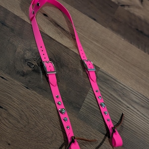 May include: A bright neon pink harness featuring two adjustable silver-toned metal buckles and a series of small, iridescent heart-shaped studs along the straps. The harness includes two loops at the bottom with thin, dark brown leather ties.