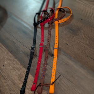 May include: Four thin, adjustable straps in black, red, brown, and orange. Each strap features a silver-toned metal buckle and a series of small, embedded circular studs along the length. The straps are arranged vertically and parallel to each other.