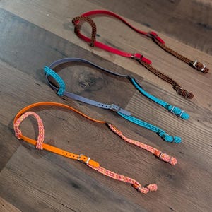 May include: Three horse halters featuring a combination of smooth synthetic straps and braided rope sections. The top halter is red and brown, the middle is light gray and turquoise, and the bottom is bright orange with a pink and orange braided rope.