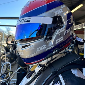 May include: A racing helmet in blue, silver, red, and white. The helmet has a blue visor and the letters "AMG" are visible. The helmet is worn by a person in a white racing suit.