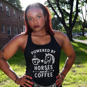 以下が含まれることがあります： コーヒーカップと馬の頭の白いスクリーン印刷グラフィックが施された黒いレーサーバックタンクトップ。胸元には「POWERED BY HORSES and COFFEE」の文字がプリントされています。シルバーカラーの多連チェーンベルトを合わせて着用しています。