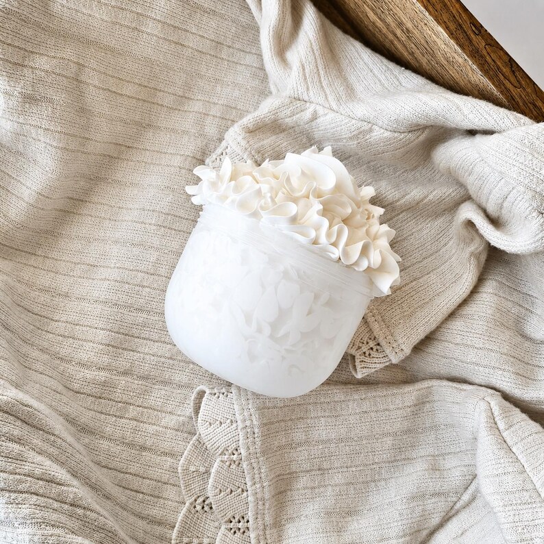 Puede incluir: Un tarro de vidrio blanco esmerilado con un dise&ntilde;o floral y una parte superior con volantes. El tarro est&aacute; lleno de material blanco con volantes y se encuentra sobre una tela beige texturizada.