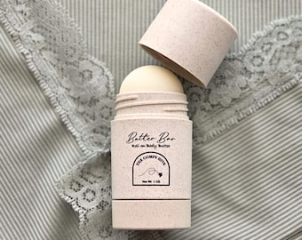 May include: A beige roll-on body butter container with the lid off, revealing the product. The container has the text "Batter Bar" and "The Comfy Hive" printed on it. The product is 28g.