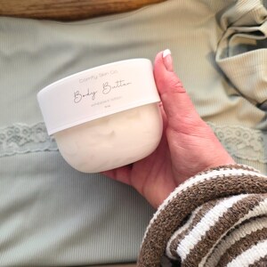 Puede incluir: Un recipiente blanco de loci&oacute;n batida "Body Butter" de Comfy Skin Co. El recipiente se sostiene en una mano, con una parte superior verde claro y una manga a rayas marrones y blancas en el fondo. La etiqueta dice "Body Butter" en cursiva.