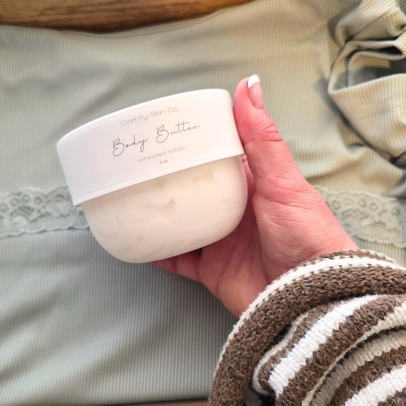 Puede incluir: Un recipiente blanco de loci&oacute;n batida "Body Butter" de Comfy Skin Co. El recipiente se sostiene en una mano, con una parte superior verde claro y una manga a rayas marrones y blancas en el fondo. La etiqueta dice "Body Butter" en cursiva.
