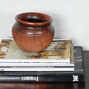 Peut inclure: Un pot en terre cuite brun est posé sur une pile de magazines. Les magazines comprennent "Milk Decoration" et "Femmes" avec le texte "dans la peau de sa grand-mère".