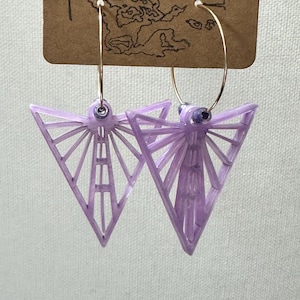 May include: A pair of lavender geometric earrings with gold-colored hoops. The earrings are triangular with a detailed, open design. The earrings are hanging from a brown tag with the text "Stellercraft".
