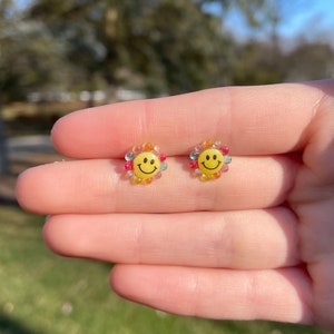 May include: A pair of yellow smiley face earrings with a colorful beaded halo. The beads are pink, blue, green, and yellow.