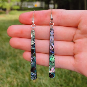 May include: A pair of long, silver dangle earrings with iridescent abalone shell accents. The earrings are a dark green, blue, and purple color.