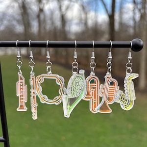 May include: A collection of musical instrument earrings, including a trumpet, flute, tambourine, and saxophone. The earrings are made of clear acrylic with colorful accents and silver-tone hooks. The instruments are suspended from a black metal bar.