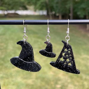 May include: Three pairs of black glitter witch hat earrings. The hats are different sizes and styles. One hat has a star design.